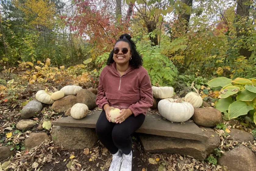 Multiracial woman smiling, wearing sunglasses sitting on a bench while holding a white gourd. Trees and blue sky in the background.