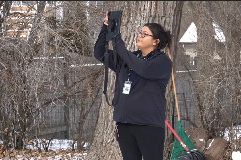 A dark-haired Lakota woman is standing outdoors. She faces to the left of the image, holding up an iPad and taking a photo. The background is snowy and filled with trees.