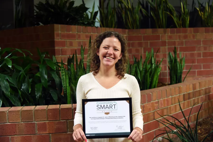 A woman stands holding up a framed certificate. She appears white with curly brown hair. SHe is smiling as she holds up the certificate that reads "Charging Smart BRONZE" and other small text that is illegible. 