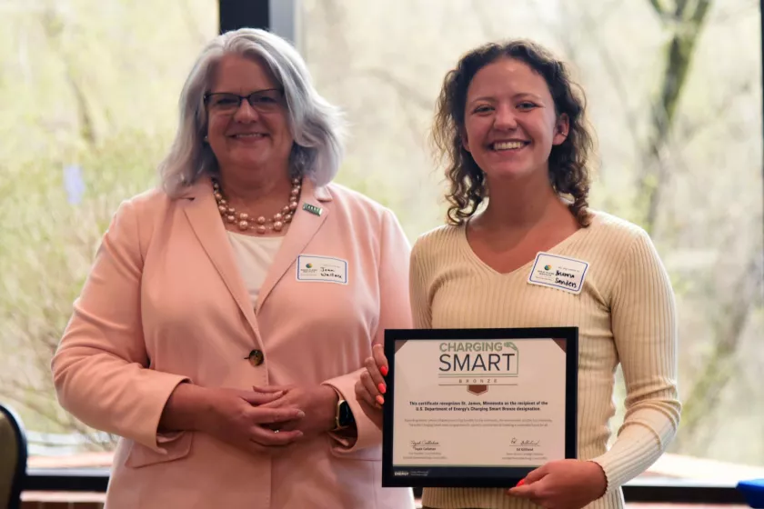 Two women pose together in front of a window. The woman on the left appears white with silver hair, she is smiling and wearing a pink blazer. The woman on the right appears white and has dark curly hair. She is holding a framed certificate and smiling. 