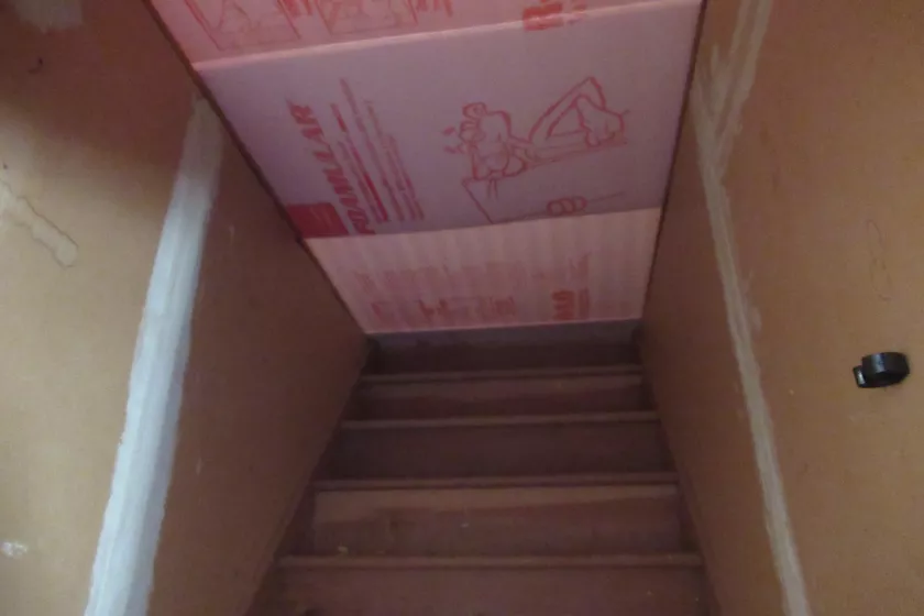 A light brown wooden staircase stretches from the bottom of the image to the middle. The top of the staircase is blocked off by a red and pink piece of foam insulation. On either side of the staircase and foam are unfinished sheetrock walls. 