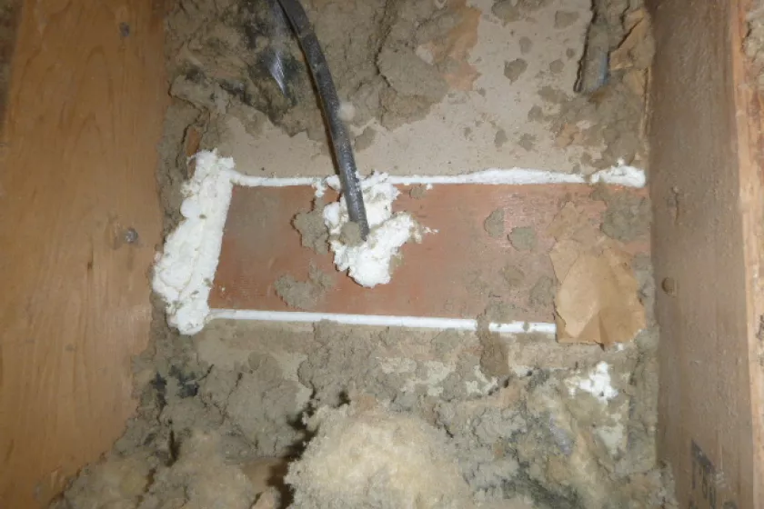 A close up shot inside the attic of a home. A black cord feeds into a piece of brown wood and the entrance point is surrounded by white foam. Around the wood is dusty insulation bits and more interior wood.