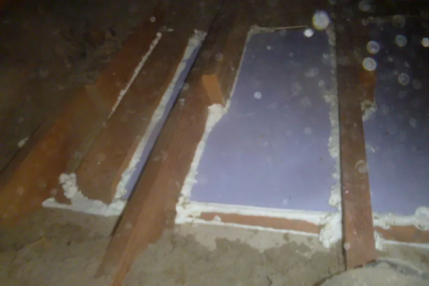 A close up shot inside the attic of a home. Three light brown wooden planks run vertically, surrounded by gray, dusty insulation fragments. Between the planks are white, flat, foam, insulation panels. Around the perimeter of the panels is white foam, sealing the edges. The image is grainy with dust particles refracting light.