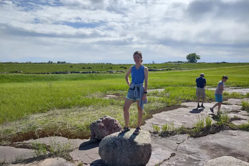 Nadya Bucklin stands on top of a rock, with bright green land stretching out behind her and a cloudy but bright sky above.