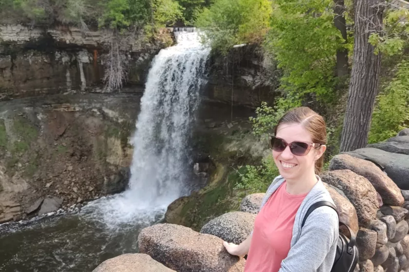 Nadya Bucklin stands smiling at the camera, behind her is a waterfall surrounded by rocky and tree-filled terrain. 