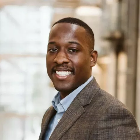 A man smiles at the camera in a professional headshot photo. He is African American and wearing a brown suit. He has short dark hair and a short mustache.  