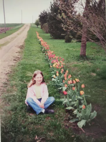 A young Nadya Bucklin sits in the grass next to a dirt road and a row of bright colorful tulips.