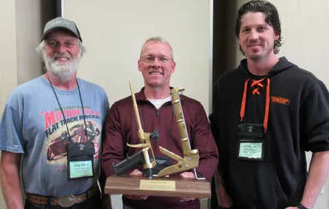 Three men standing shoulder to shoulder smiling. In his hands, the middle man holds a trophy. The trophy has a solid wood base with gold foam gun and a gold caulk gun displayed.