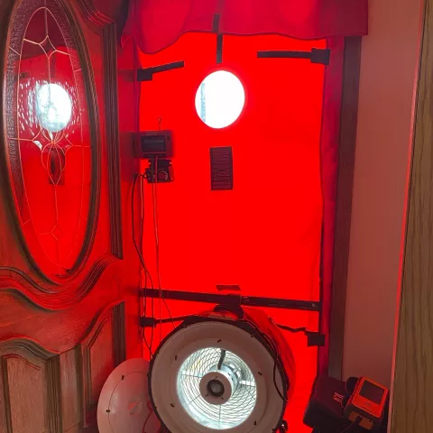 A foyer inside a home. A brown wooden front door with a large oval window is propped open. A large red piece of plastic is covering the doorway. The cover is casting a red glare throughout the foyer. Near the floor, there is an opening in the cover that holds a circular fan. To the right of the door sits weatherization electrical equipment.