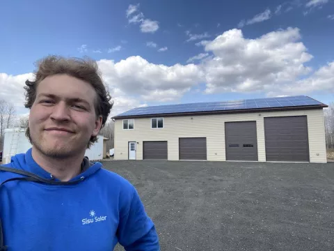 A young man poses for a selfie outdoors. He is a white man with blonde hair, wearing a blue hoodie with the logo for Sisu Solar on the chest. He is smiling at the camera. Behind him stands a four door garage with solar panels mounted on the roof. The weather appears slightly overcast but blue skies can be seen in the background. 