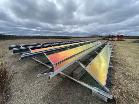 Solar panels stand in on open land. The panels reflect a rainbow light and the surrounding land is seasonally brown. Above sits a overcast sky with rolling white clouds.