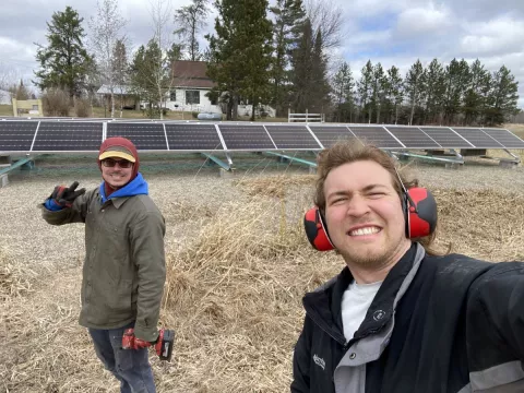 Two smiling men stand in front of solar panels outdoors. The man on the left is wearing a hooded coat, sunglasses and gloves. In one hand he holds a drill and with the old he flashes a peace sign toward the camera. The man on the right is taking the selfie and wearing large protective headphones.