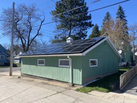 A detached garage with a solar panel mounted on the roof. The corresponding home can be seen in the background, both buildings are painted mint green with white trim.