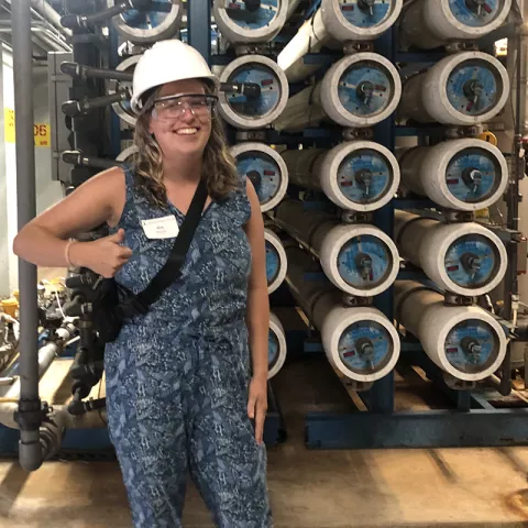 White woman with light hair wearing a white hard hat and holding her thumb up. In the background is a mechanical warehouse environment. 