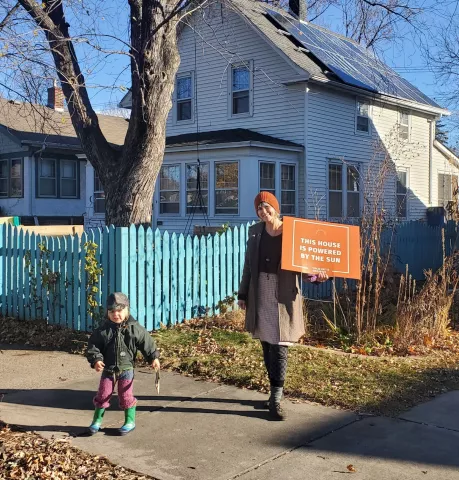 Woman and child standing outside with a house and garden in the background. Woman is holding an orange sign that reads "THIS HOUSE IS POWERED BY THE SUN."