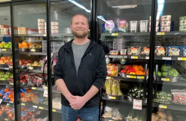 Bearded white man stands in front of a floor to ceiling cooler. Cooler is filled with fresh fruits and vegetables.