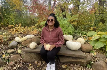 Multiracial woman wearing sunglasses sitting on a bench while holding a white gourd. Trees and blue sky in the background.