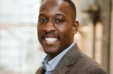 A man smiles at the camera in a professional headshot photo. He is African American and wearing a brown suit. He has short dark hair and a short mustache.