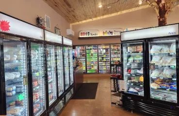 The inside on a convenience store. Three large, standup coolers can be seen. They shine bright white light and are stocked with items like pop, bags of frozen food, and other grocery items. The ceiling, walls and floor and all warm shades of brown and look freshly constructed. A large, decorative birch tree trunk and bare limbs can be seen above the coolers, reaching toward the ceiling. 