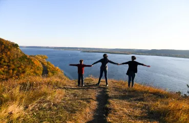 A yellow grassy knoll overlooking a bright blue body of water and a clear blue sky above. On the knoll stands three people, an adult and two children, with their arms stretched horizontally outward. Their features can't be made out but they appear to be looking toward the water. 