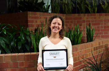 A woman stands holding up a framed certificate. She appears white with curly brown hair. SHe is smiling as she holds up the certificate that reads "Charging Smart BRONZE" and other small text that is illegible. 