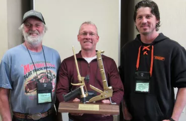  Three men standing shoulder to shoulder smiling. In his hands, the middle man holds a trophy. The trophy has a solid wood base with gold foam gun and a gold caulk gun displayed.