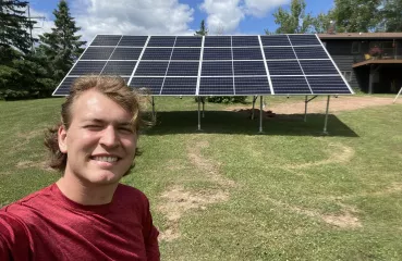 A young man takes an outdoor selfie. In the foreground, the white, blonde man wear a red shirt and squints into the sun with a big smile. Behind him a large solar panel stands in a grassy area.