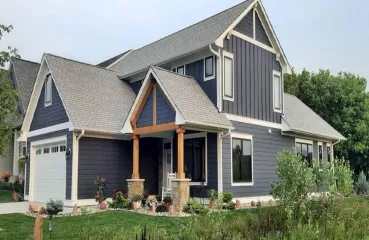 A large blue home with a gray roof and white trim. There is green grass in the foreground and the background shows green trees and blue skies. 