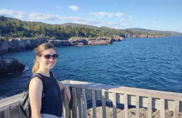 Nadya Bucklin stands on a deck overlooking a large blue body of water with green trees and rocky shores visable. 