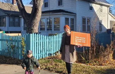 Woman and child standing outside with a house and garden in the background. Woman is holding an orange sign that reads "THIS HOUSE IS POWERED BY THE SON."