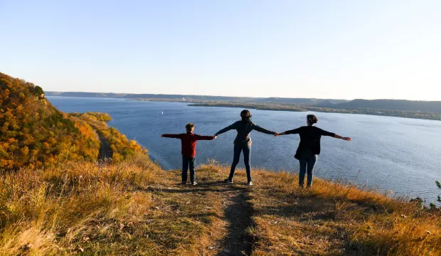 A yellow grassy knoll overlooking a bright blue body of water and a clear blue sky above. On the knoll stands three people, an adult and two children, with their arms stretched horizontally outward. Their features can't be made out but they appear to be looking toward the water. 