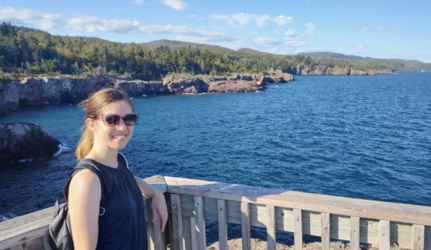 Nadya Bucklin stands on a deck overlooking a large blue body of water with green trees and rocky shores visable. 